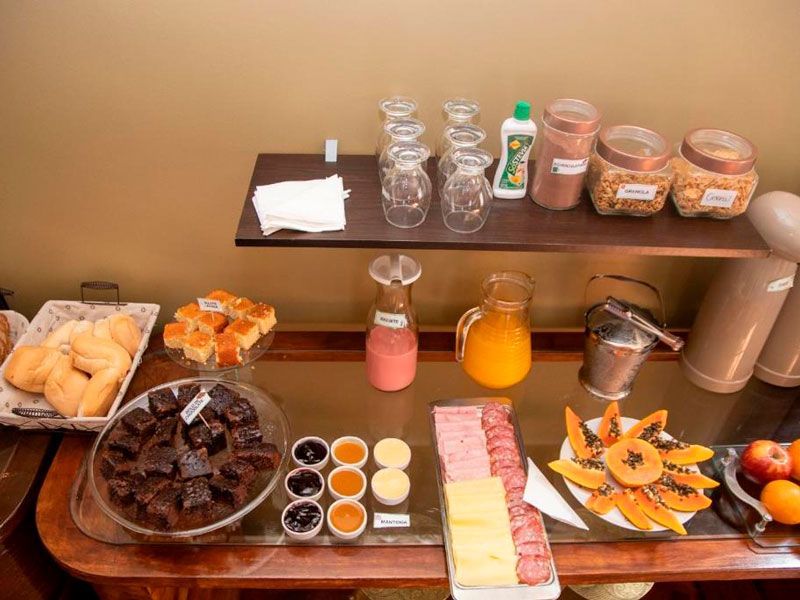 Um buffet de café da manhã com doces, frutas, frios, queijos, geleias, copos e jarras dispostos em prateleiras e em uma mesa.