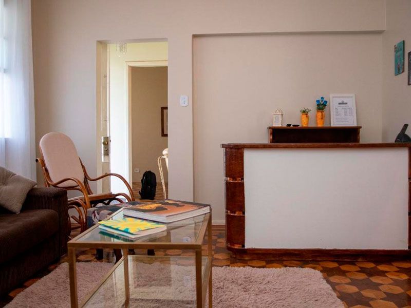 Uma área de espera com uma mesa de centro de vidro, uma cadeira de vime e um balcão de recepção encostado em uma parede de cor clara.