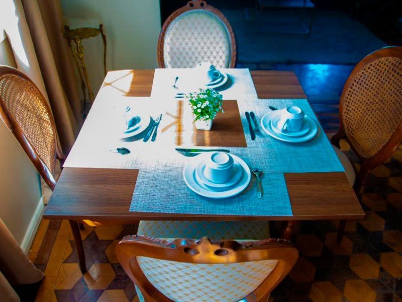 Conjunto de mesa de jantar quadrada de madeira para três pessoas, com jogos americanos azuis, xícaras e pires brancos e uma pequena planta no centro.