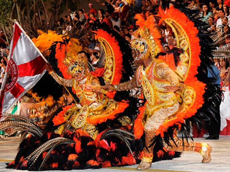 Duas artistas com trajes ornamentados e vibrantes em tons de laranja e dourado, adornados com penas, dançam em um carnaval, uma delas acenando com uma bandeira.