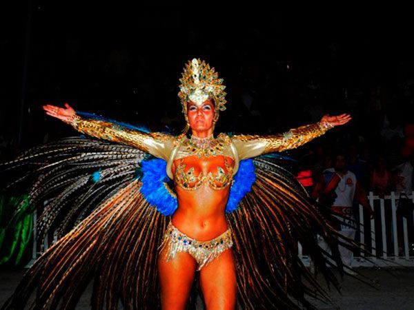 Uma pessoa vestida com uma grande fantasia de carnaval, repleta de penas, detalhes dourados e uma coroa, permanece em pé com os braços estendidos à noite.