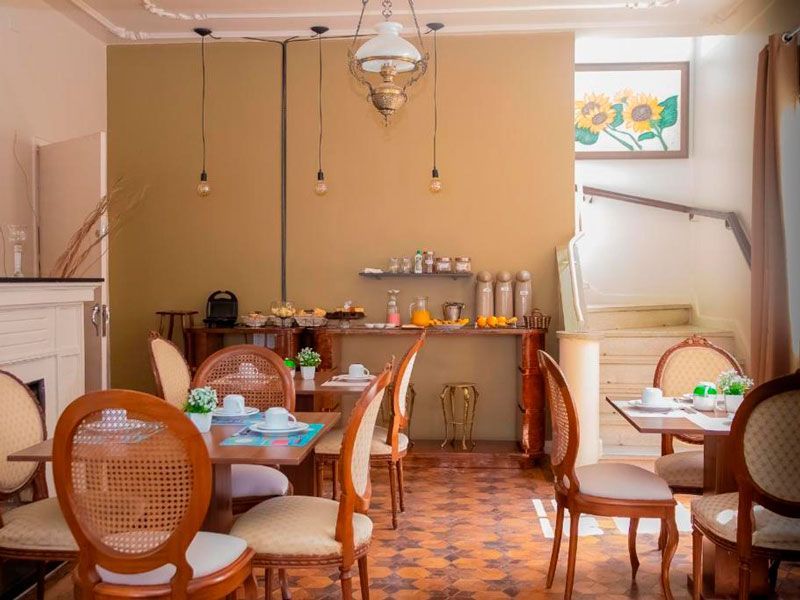 Uma sala de jantar com mesas de madeira, cadeiras com encosto de palhinha, paredes em tons quentes, luminárias pendentes e um quadro de girassóis.