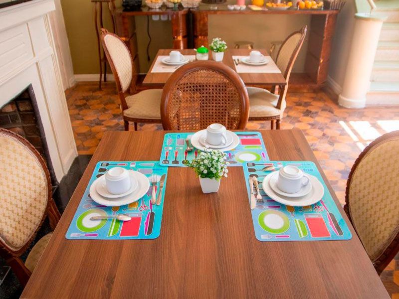 Uma mesa de jantar de madeira para duas pessoas com jogos americanos coloridos, pratos brancos, canecas e pequenos vasos de plantas em uma sala iluminada.