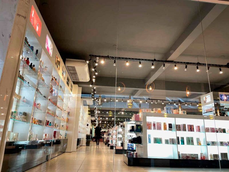 Interior de uma loja com prateleiras bem iluminadas exibindo frascos de perfume, com luzes penduradas e uma pessoa caminhando ao fundo.