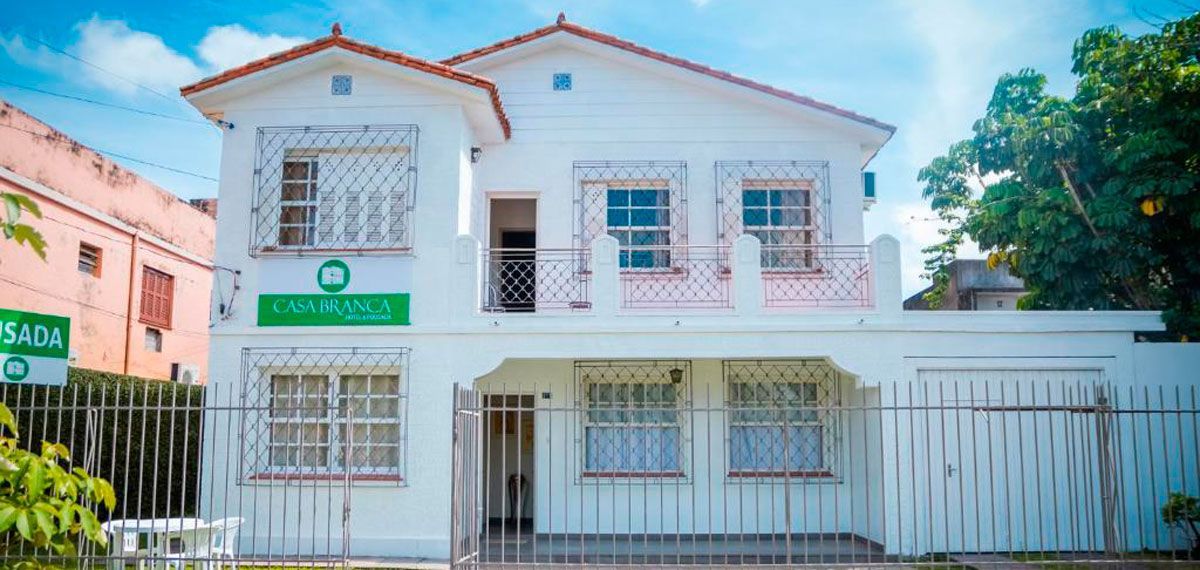 Uma casa branca de dois andares com um quintal frontal cercado, placa verde e uma varanda em um dia ensolarado.