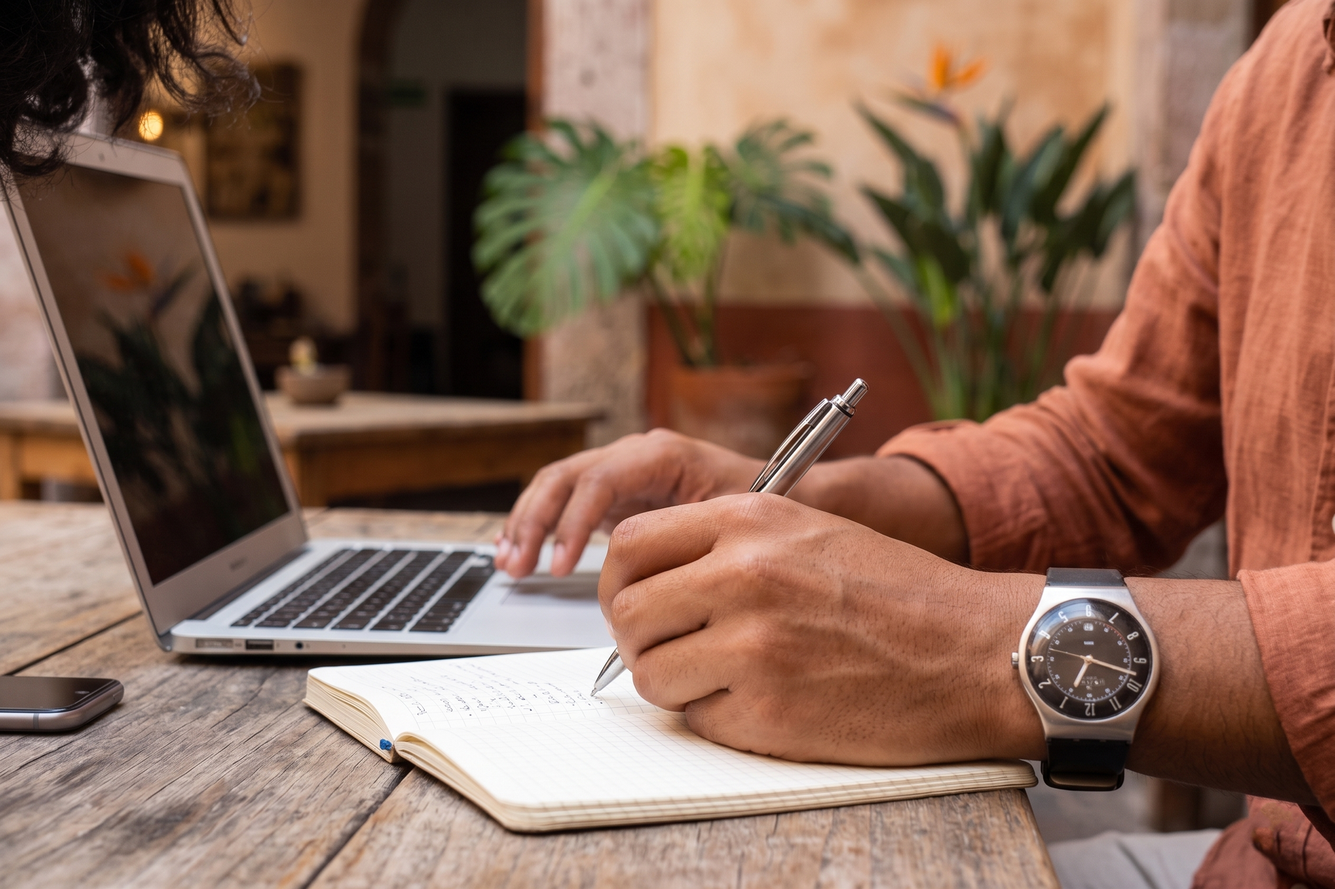 Person writing in a notebook beside a laptop at a wooden table with plants in the background