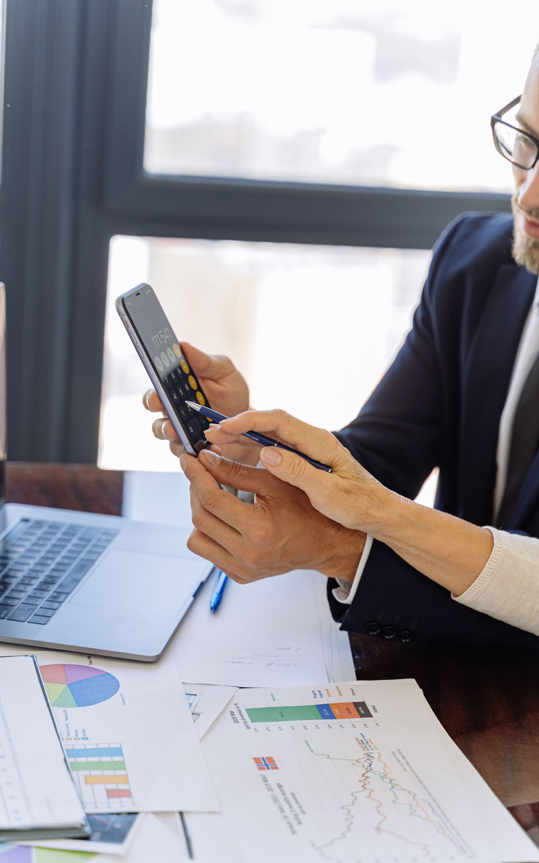 Person using a smartphone at a desk with laptop, documents, and charts in a bright office