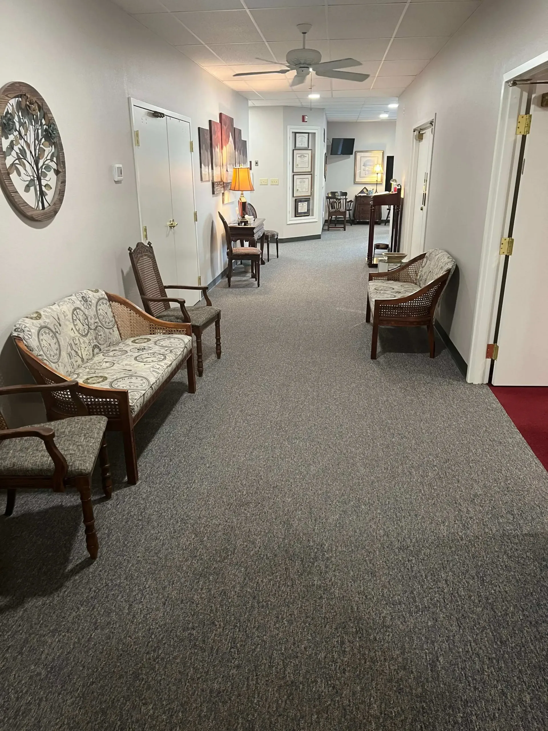 Narrow carpeted hallway with wooden chairs, wall clock, and white doors in a quiet indoor waiting area