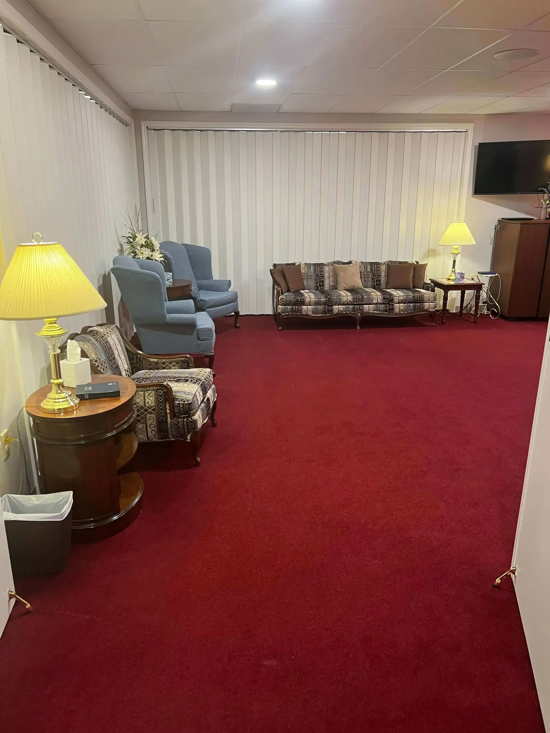 Red-carpeted lounge with blue and floral chairs, side tables, and a curtained wall at the far end.
