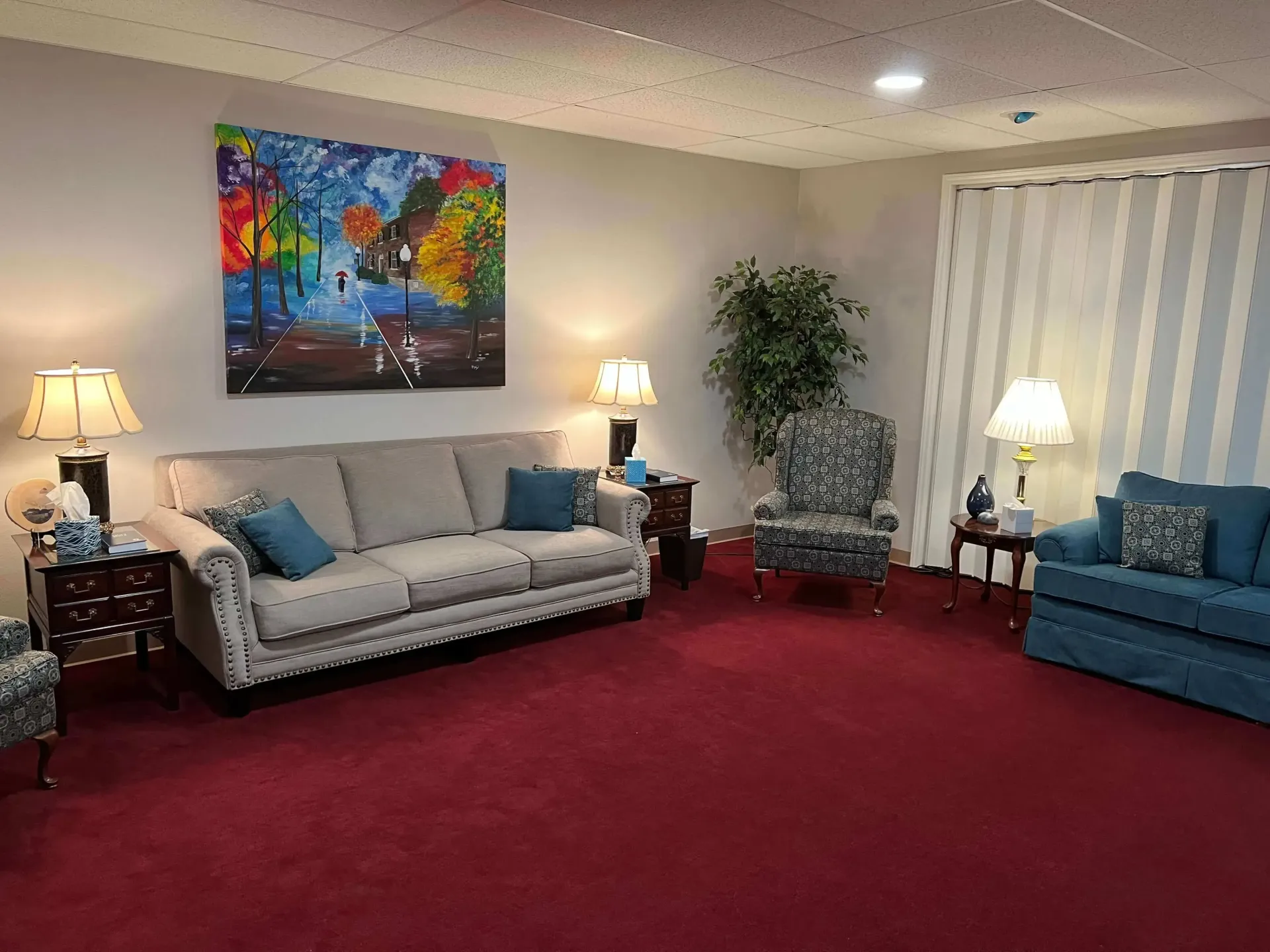 Cozy living room with red carpet, gray and blue sofas, lamps, and a colorful painting on the wall