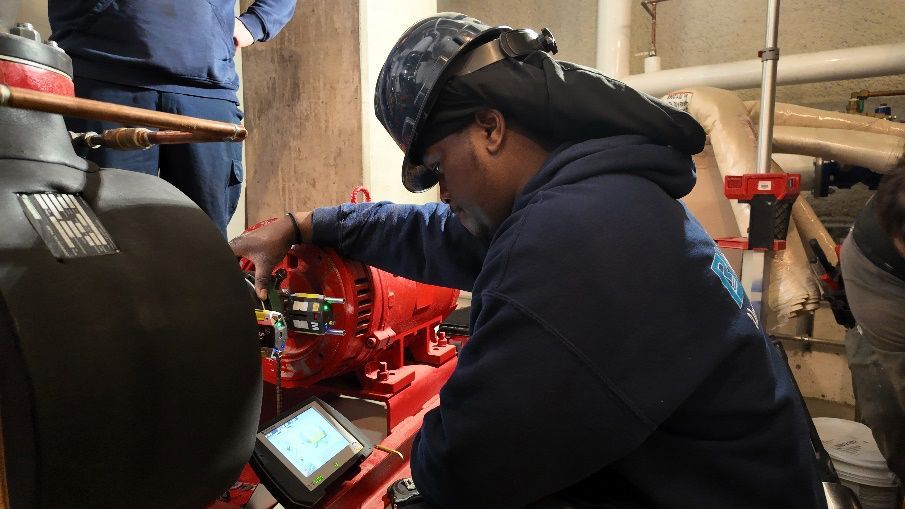 Une personne portant un casque de soudeur travaille sur une pompe rouge, à l'aide d'un appareil portatif, dans un local technique.