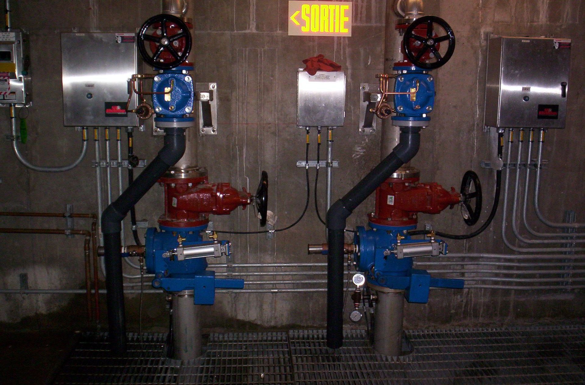 Deux vannes industrielles bleues et rouges avec des tuyaux noirs et des boîtes électriques sur un mur en béton.