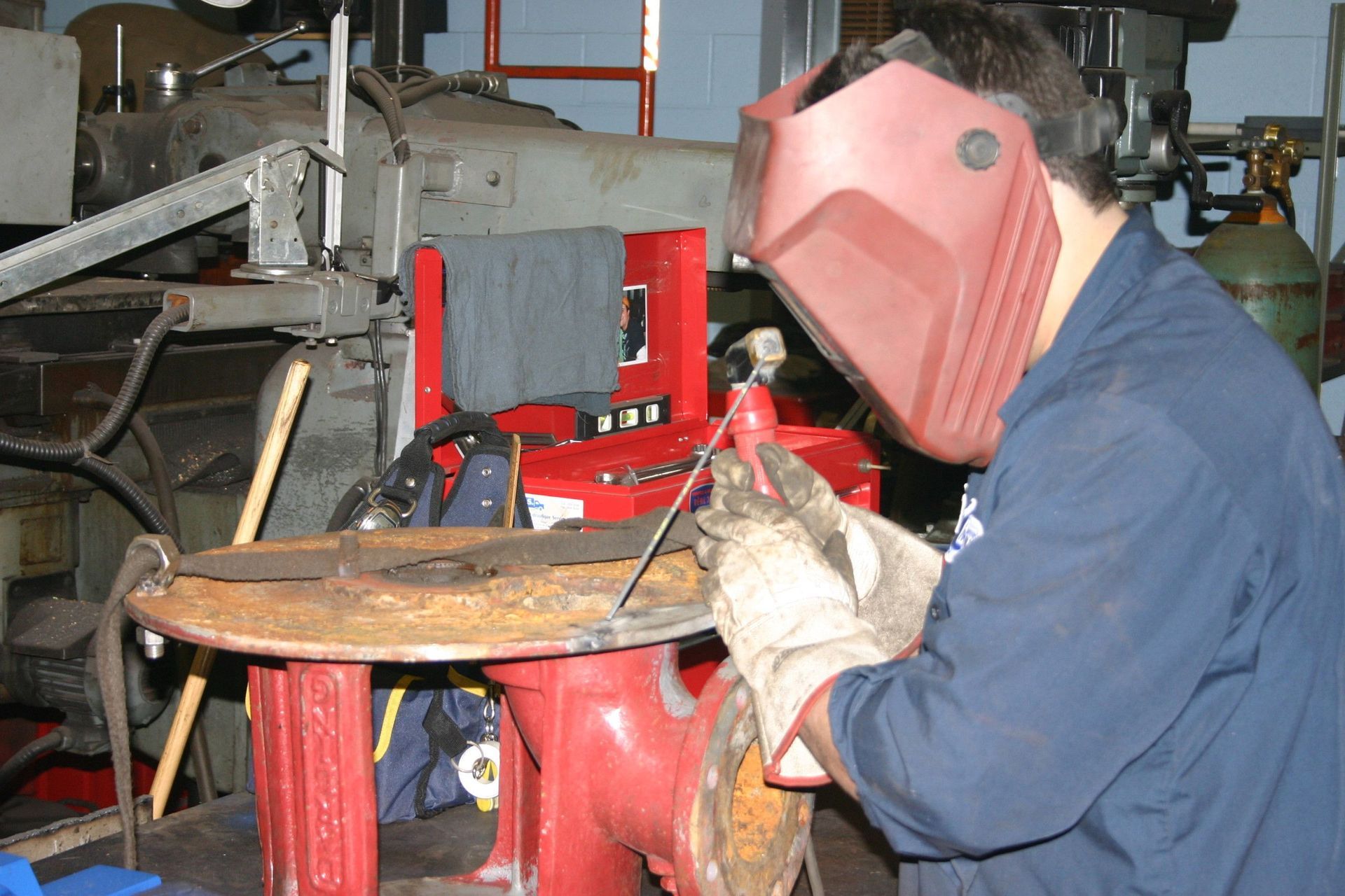 Un soudeur portant un masque rouge, des gants et une combinaison bleue travaille sur du métal dans un atelier, près de machines.