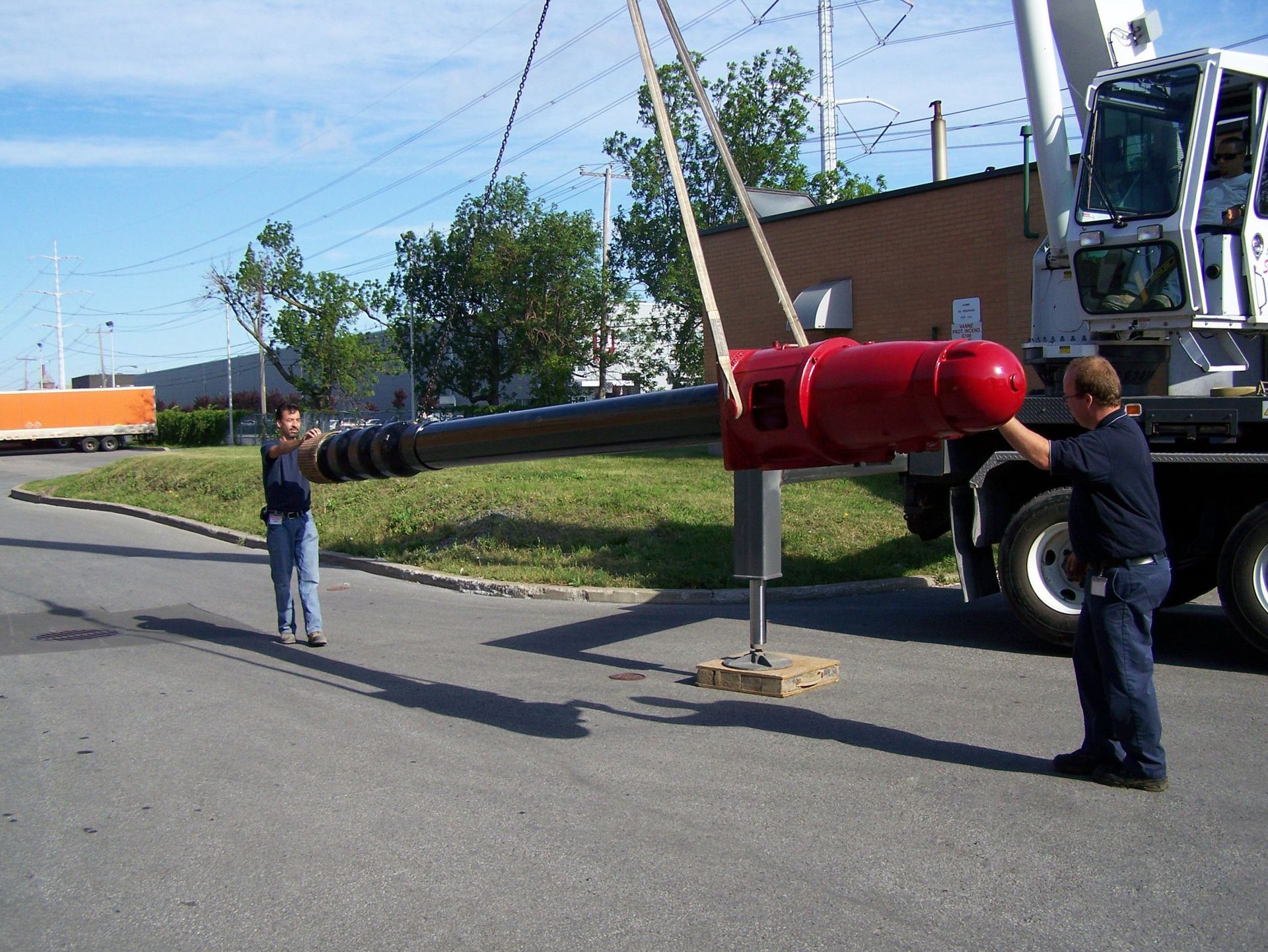 Deux hommes manœuvrant une longue tarière noire à moteur rouge, suspendue par une grue. En extérieur, sur le trottoir.