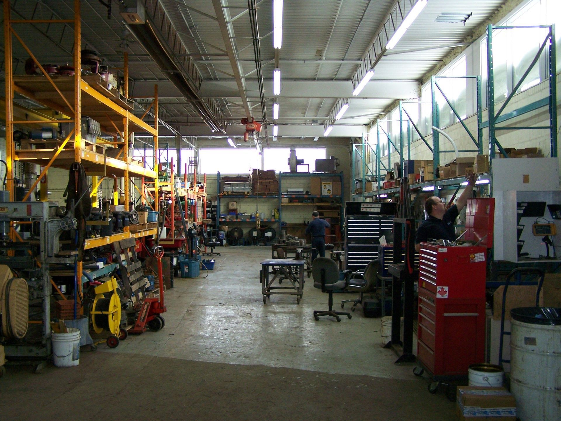 Dans un atelier industriel encombré : des étagères remplies d'outils, une boîte à outils rouge et des ouvriers près des machines.