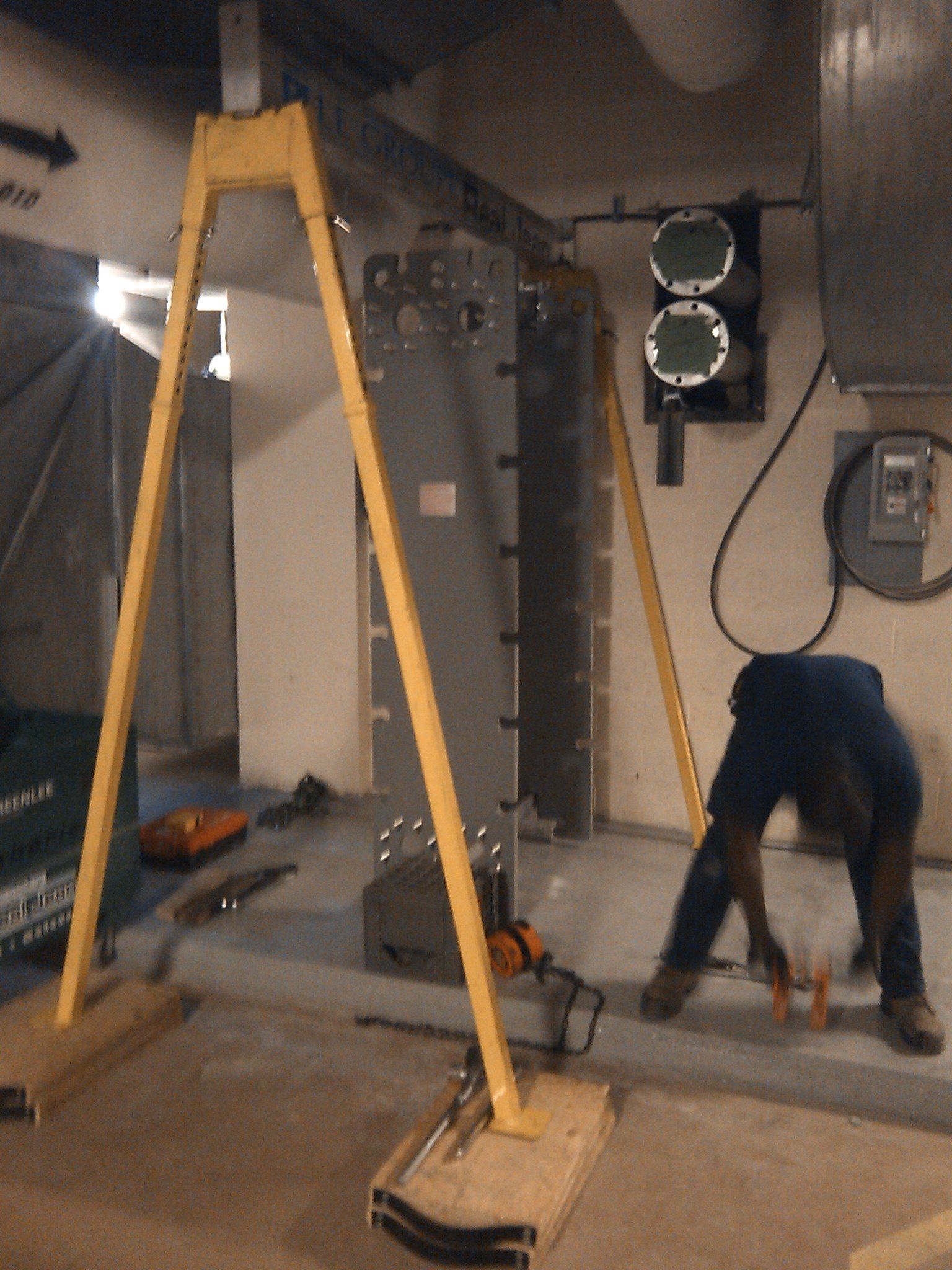 Un ouvrier se trouve près d'un grand panneau vertical gris soutenu par un cadre en A jaune. À l'intérieur, on aperçoit du matériel en arrière-plan.