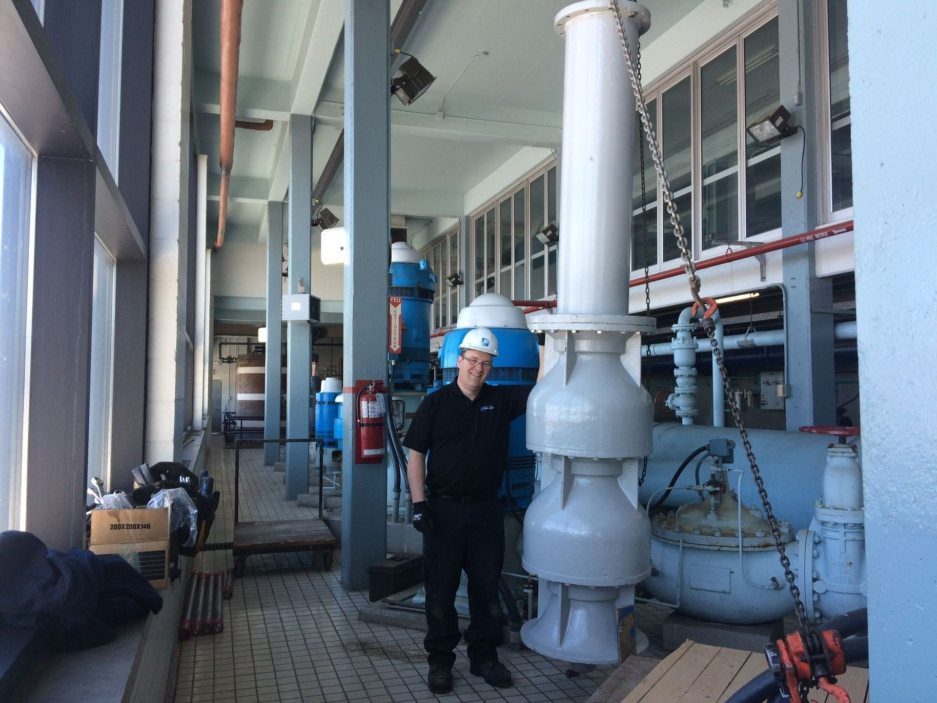 Un homme portant un casque de chantier se tient dans un intérieur industriel avec de grandes machines bleues et blanches ; des fenêtres sont visibles à gauche.
