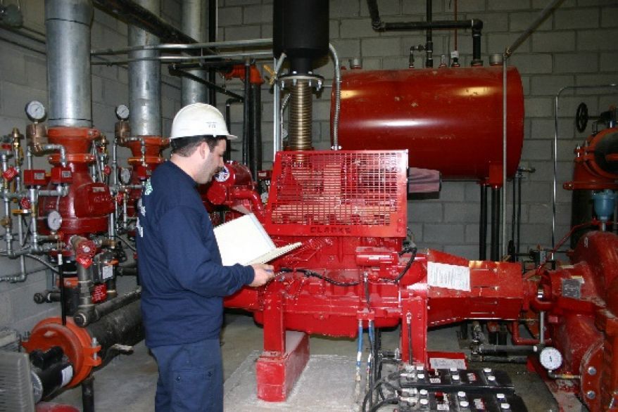 Une personne portant un casque de chantier inspecte des équipements industriels rouges, composés de tuyaux, dans un local technique, en tenant un bloc-notes.