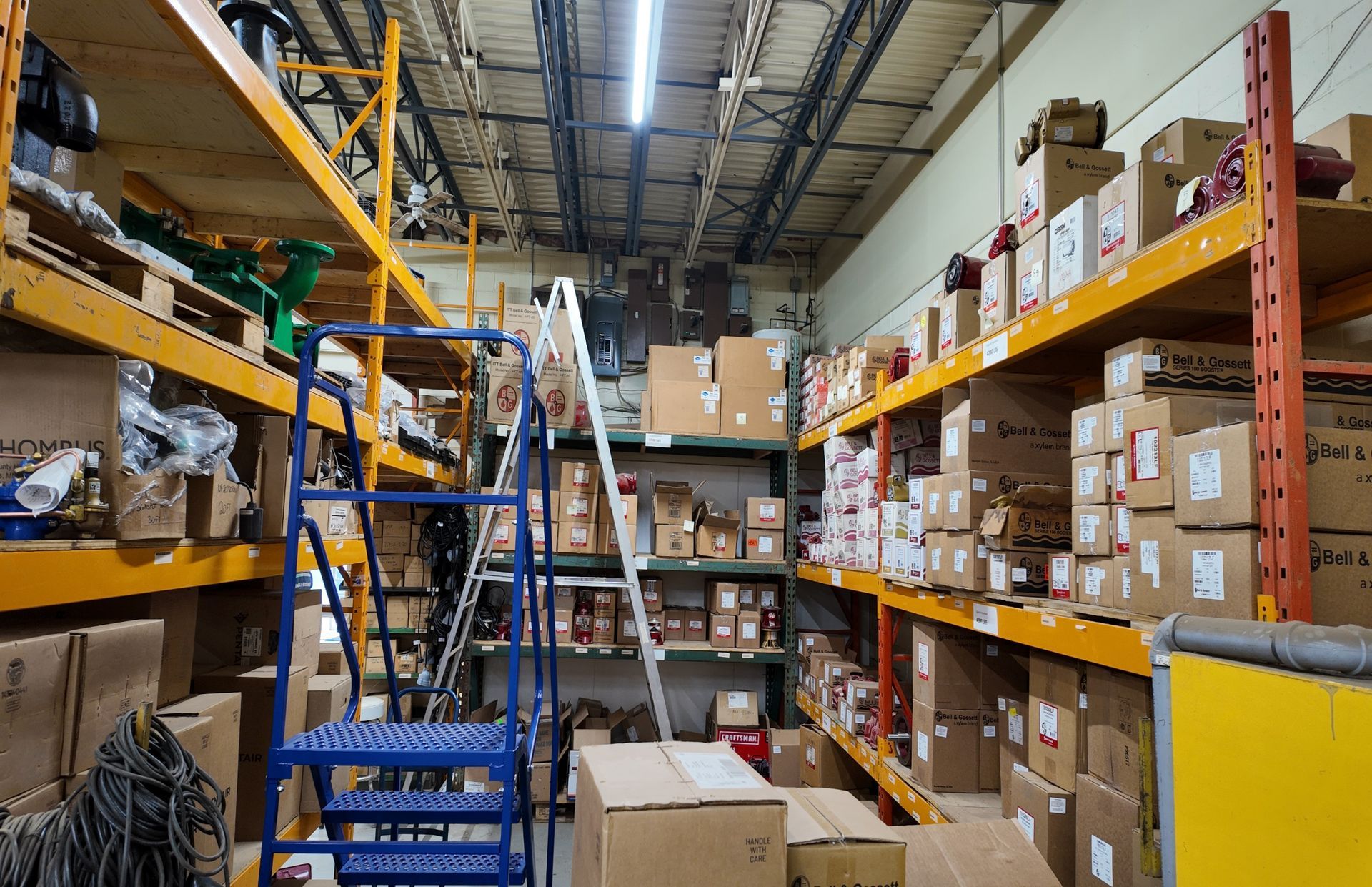 Intérieur d'un entrepôt avec de hautes étagères remplies de cartons et d'équipements ; un escabeau bleu se trouve dans l'allée.