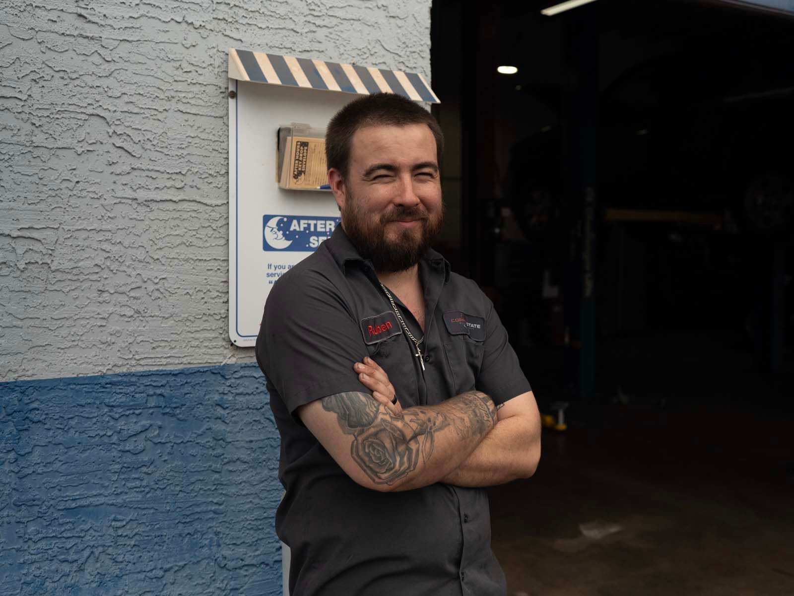Man with crossed arms and tattooed forearms standing beside a blue and gray wall near an open doorway