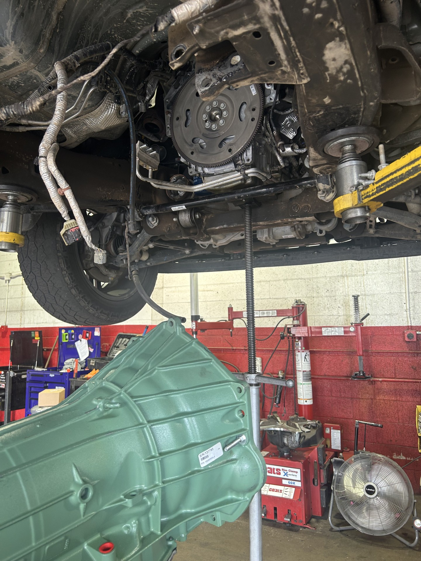Car lifted in a garage with transmission removed; green transmission jack below and tools in the background