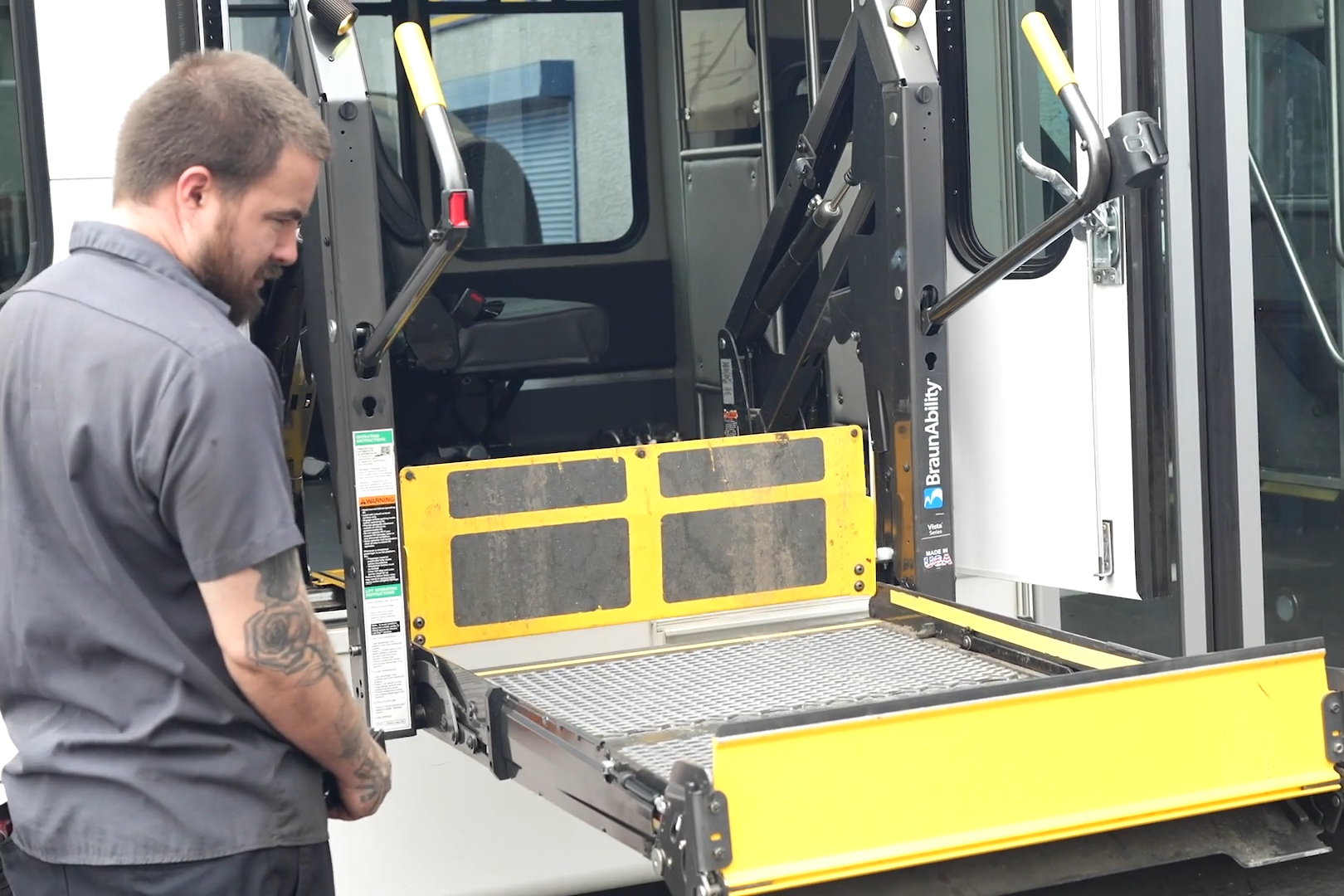 Man loading a wheelchair lift platform at the back of a bus.
