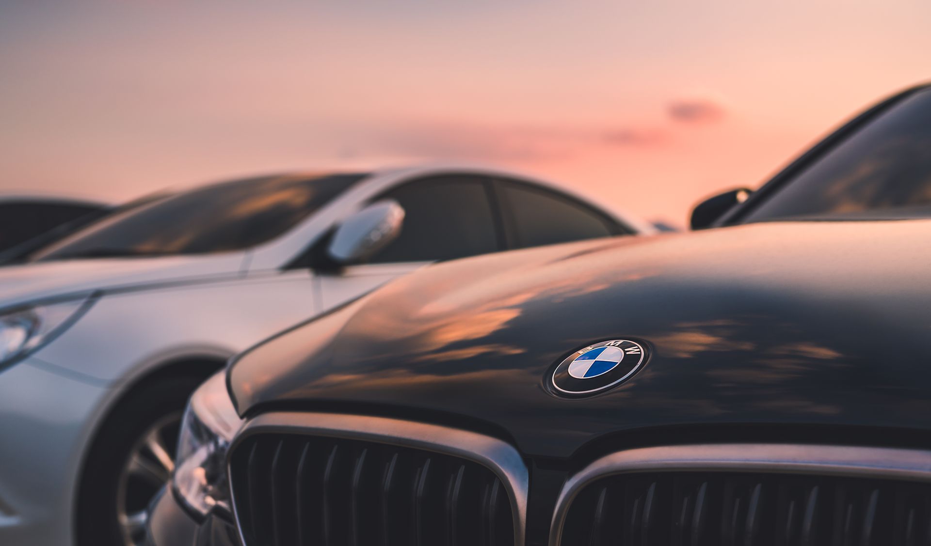Two BMW cars parked side by side at sunset, one black in the foreground and one white behind it