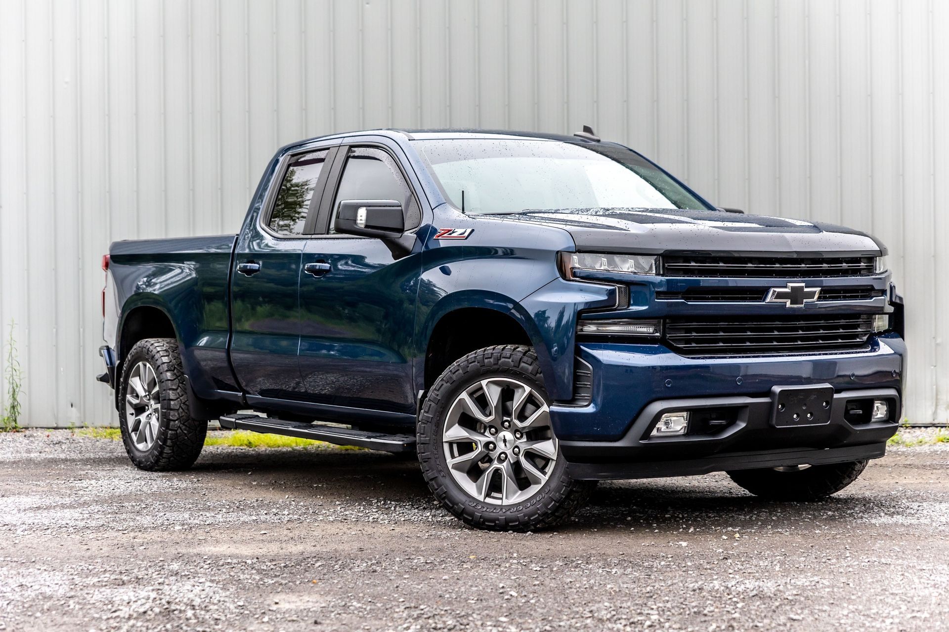 Blue Chevrolet pickup truck parked on wet pavement in front of a light gray metal wall