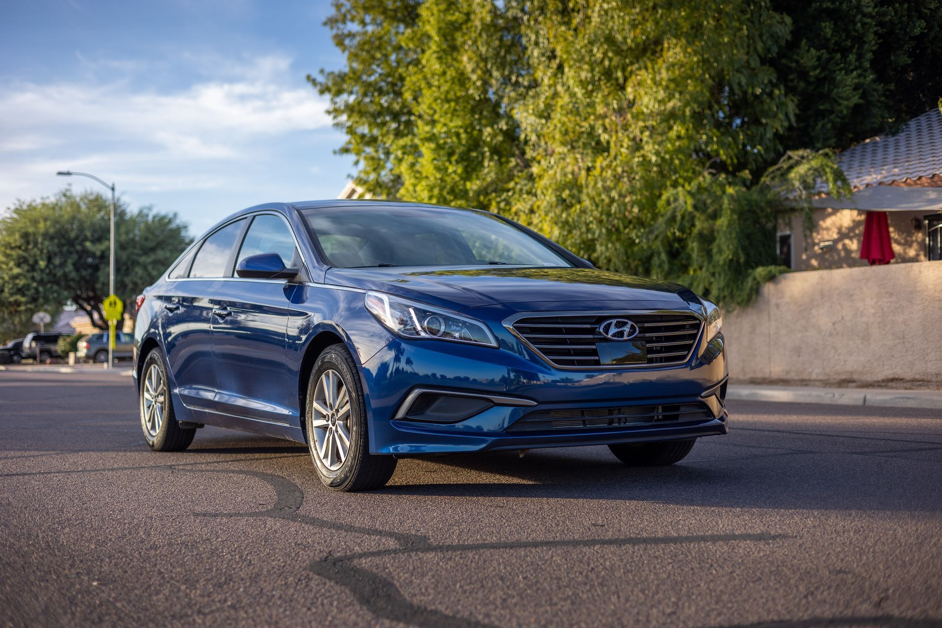 Blue sedan driving on a sunlit road with trees and a wall in the background