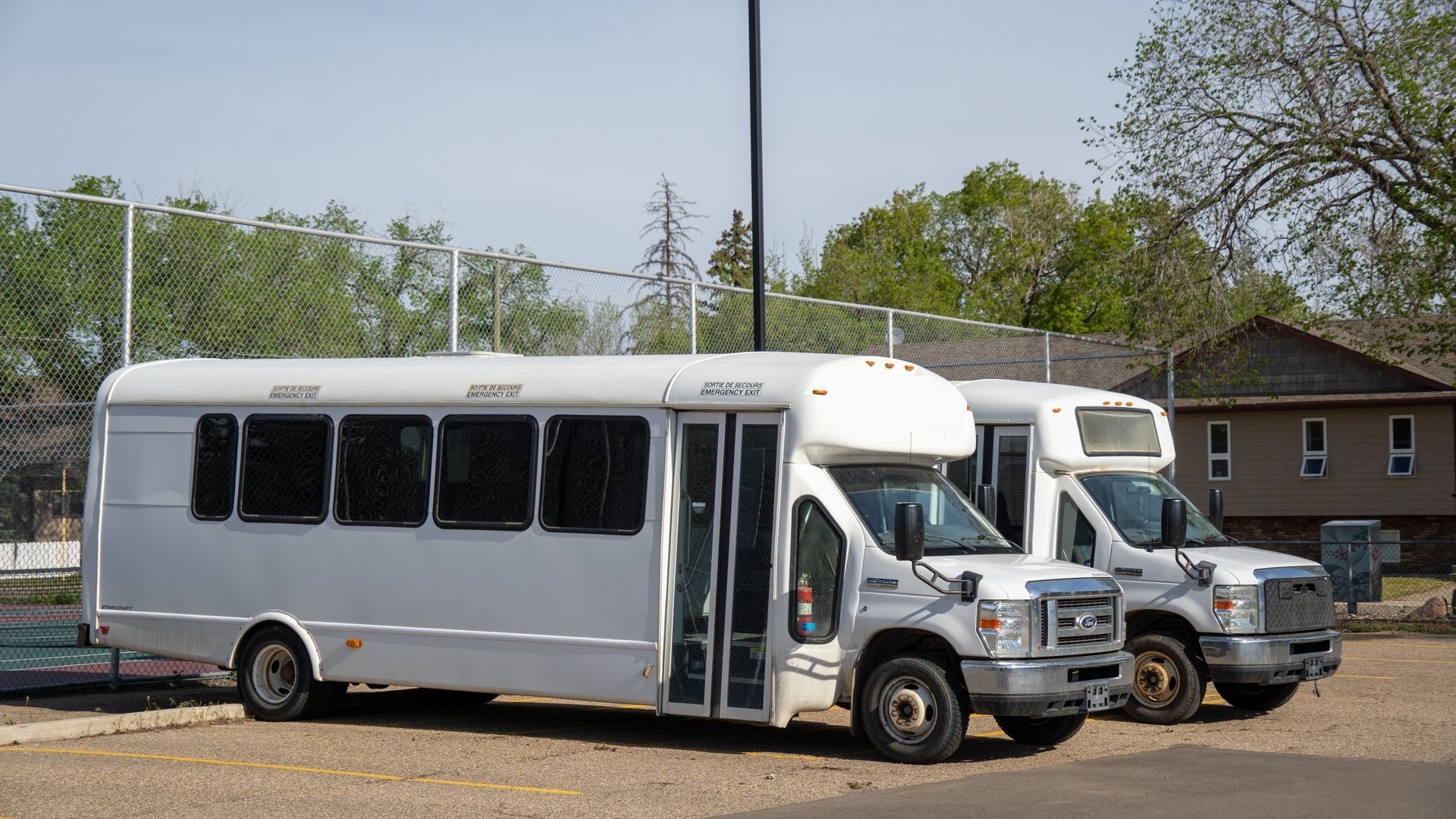 Two white shuttle buses parked side by side in a lot, with a building and trees in the background.
