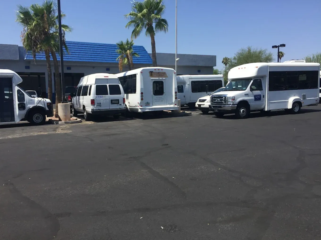 Parking lot with white vans and a shuttle bus parked near a blue-roofed building and palm trees.