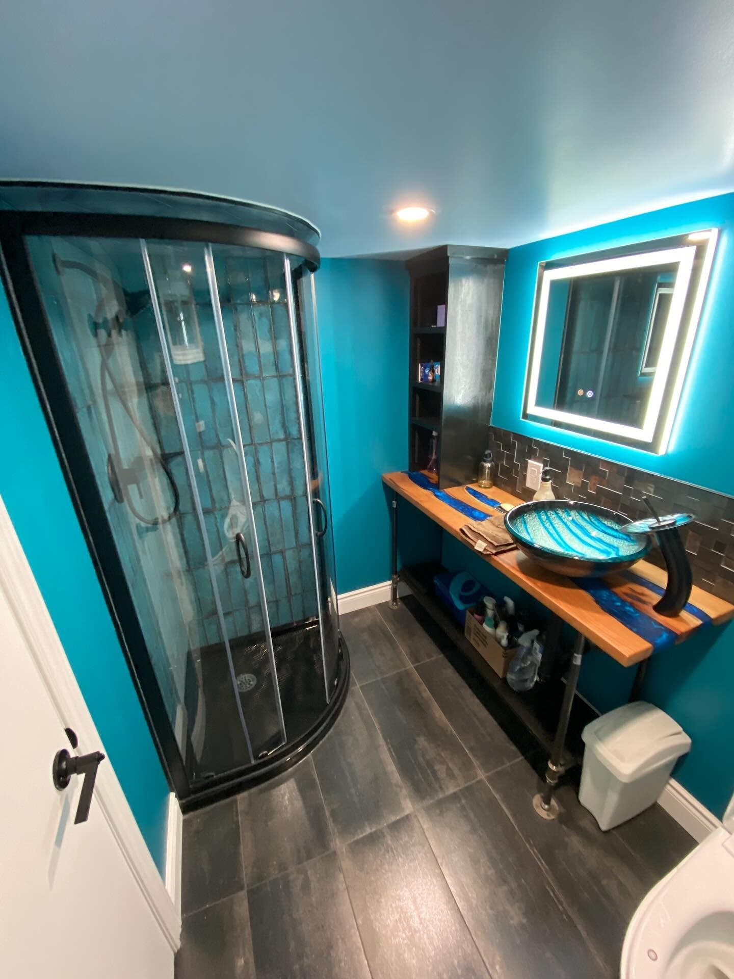Modern bathroom with curved glass shower, blue walls, wood vanity, and lit mirror.