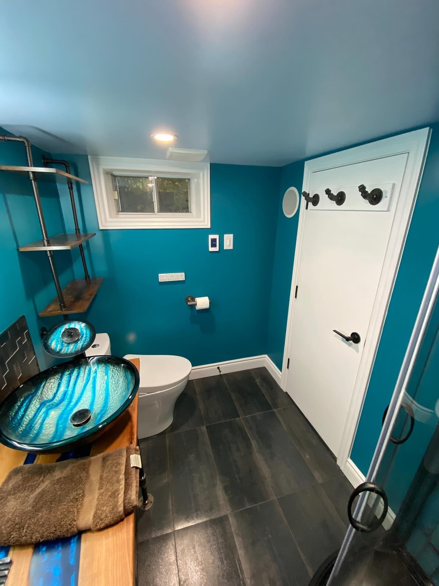 Blue bathroom with toilet, white door, small window, and glass sink on a wooden counter