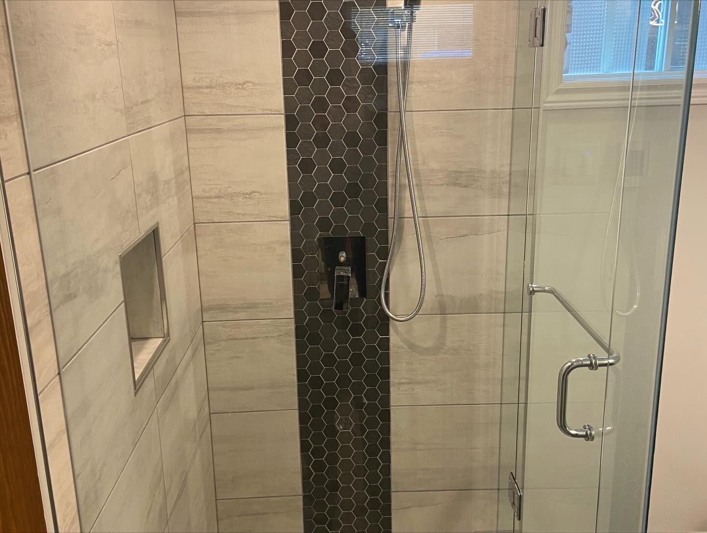 Modern shower with beige tile, dark accent wall, glass door, and handheld showerhead.