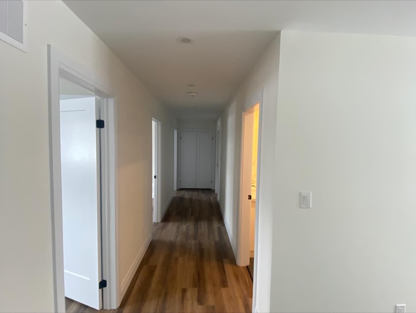 Long white hallway with wood floors and open doors, lit by natural light at the far end