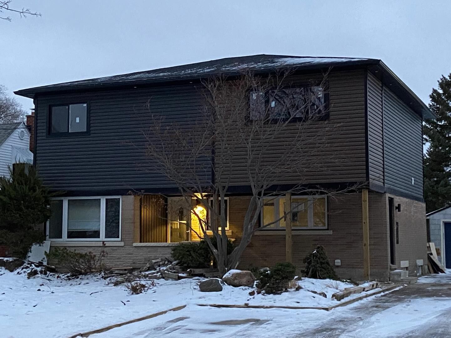 Two-story house with dark siding and lit porch at dusk, surrounded by snow and leafless trees.