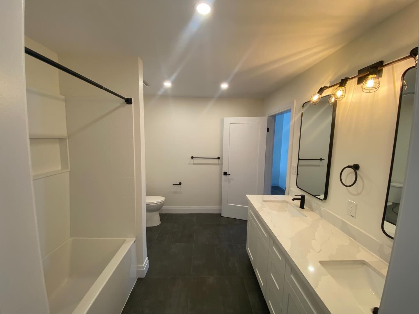 Bright modern bathroom with white vanity, mirrors, tub, toilet, and dark floor tiles