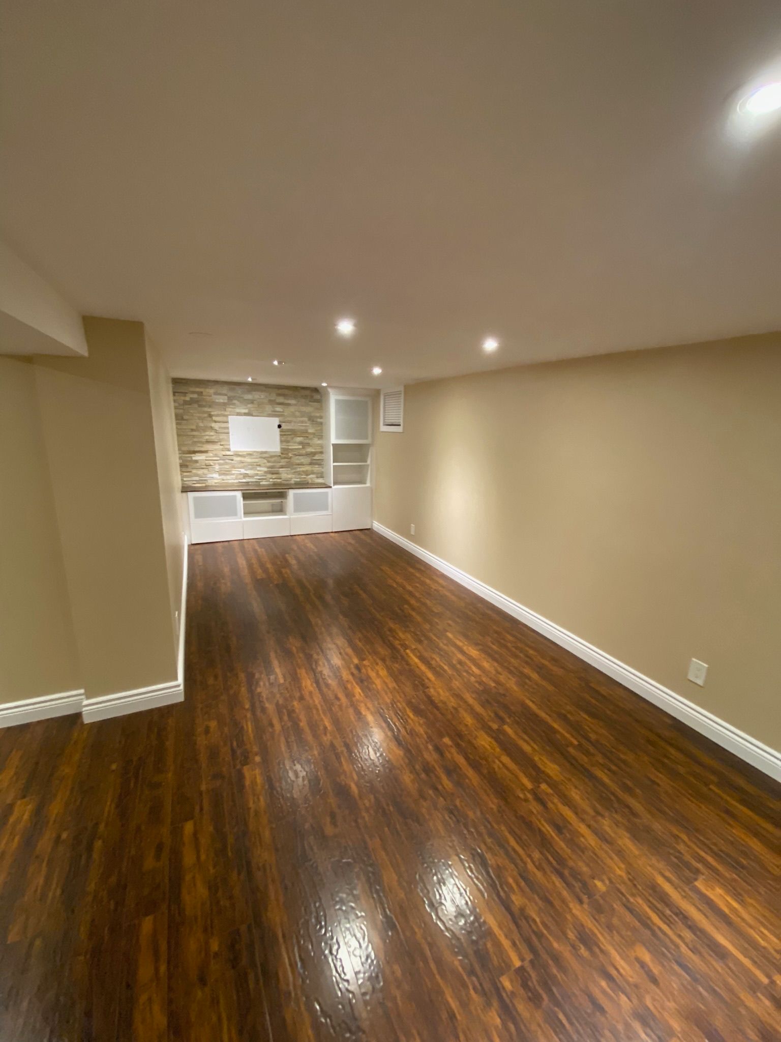 Empty finished basement with dark wood floors, beige walls, recessed lights, and a stone accent wall.