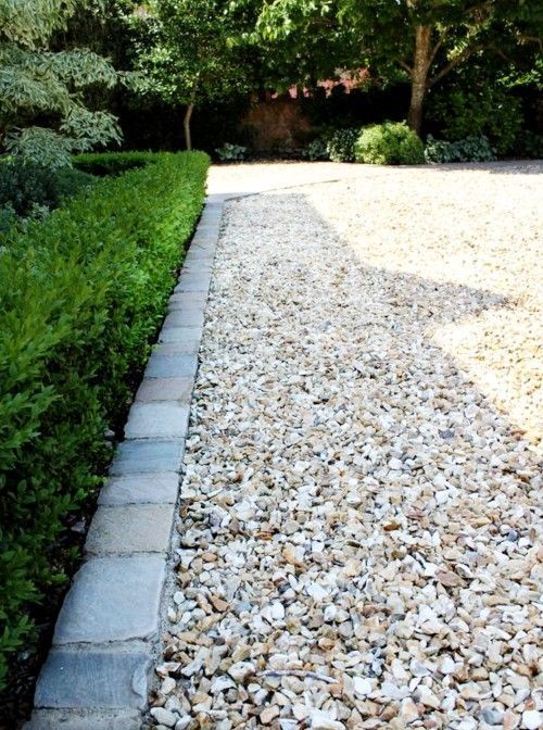 Gravel path bordered by gray bricks and a green hedge, leading towards trees and a driveway.