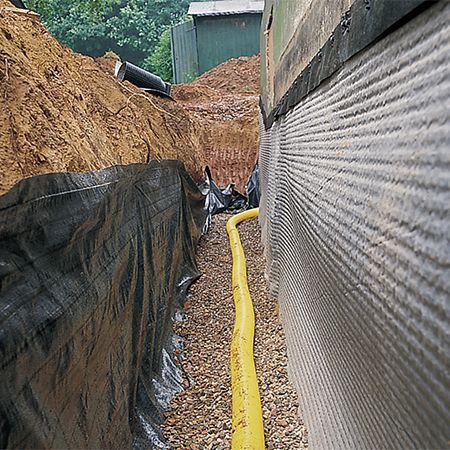 A trench next to a building foundation with drainage pipe and gravel, covered by black fabric and soil.