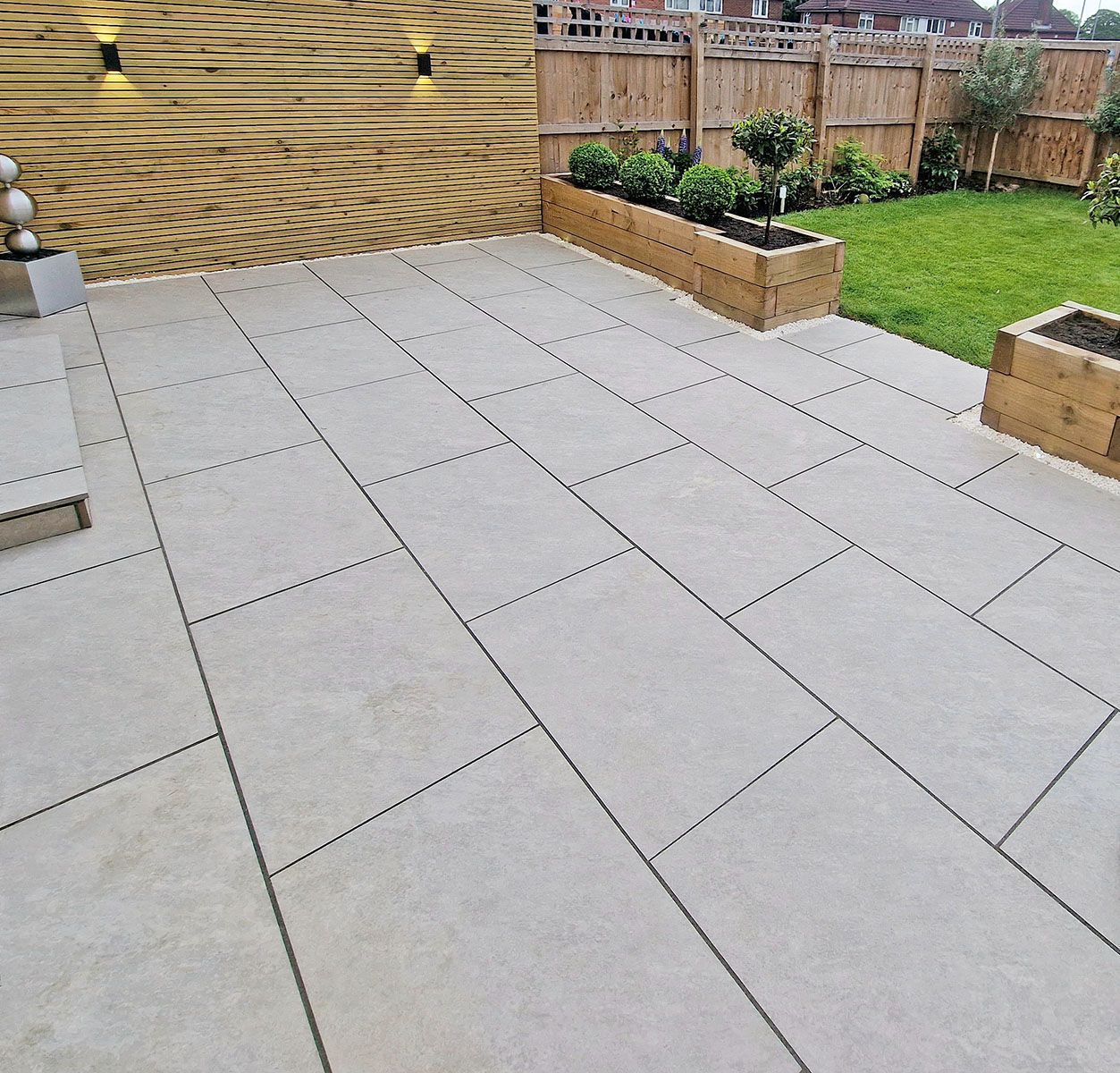 Beige stone patio with a wooden fence, raised flower beds, and a small lawn.