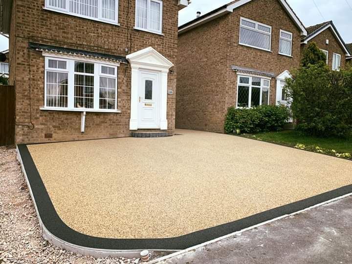 Beige resin driveway with black border in front of a brick house.