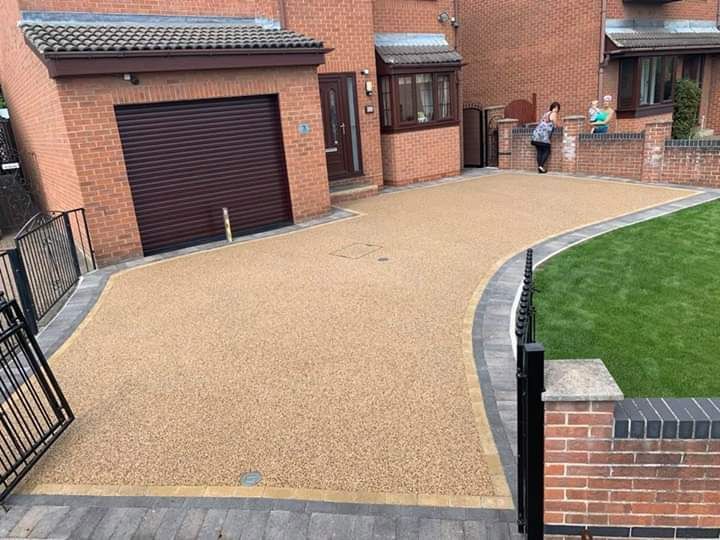 Driveway with tan resin surface, brick edging, and a green lawn. Two people stand near the house.