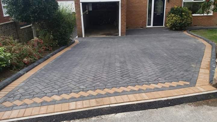 Brick driveway with brown edging, leading to a garage. Green bushes and grass on sides.