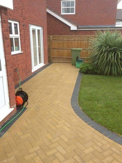 Brick patio with grass border next to a red brick building, with a garden in the background.