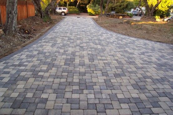 Brick-paved driveway curving towards a background with parked vehicles and trees; stones in neutral colors.