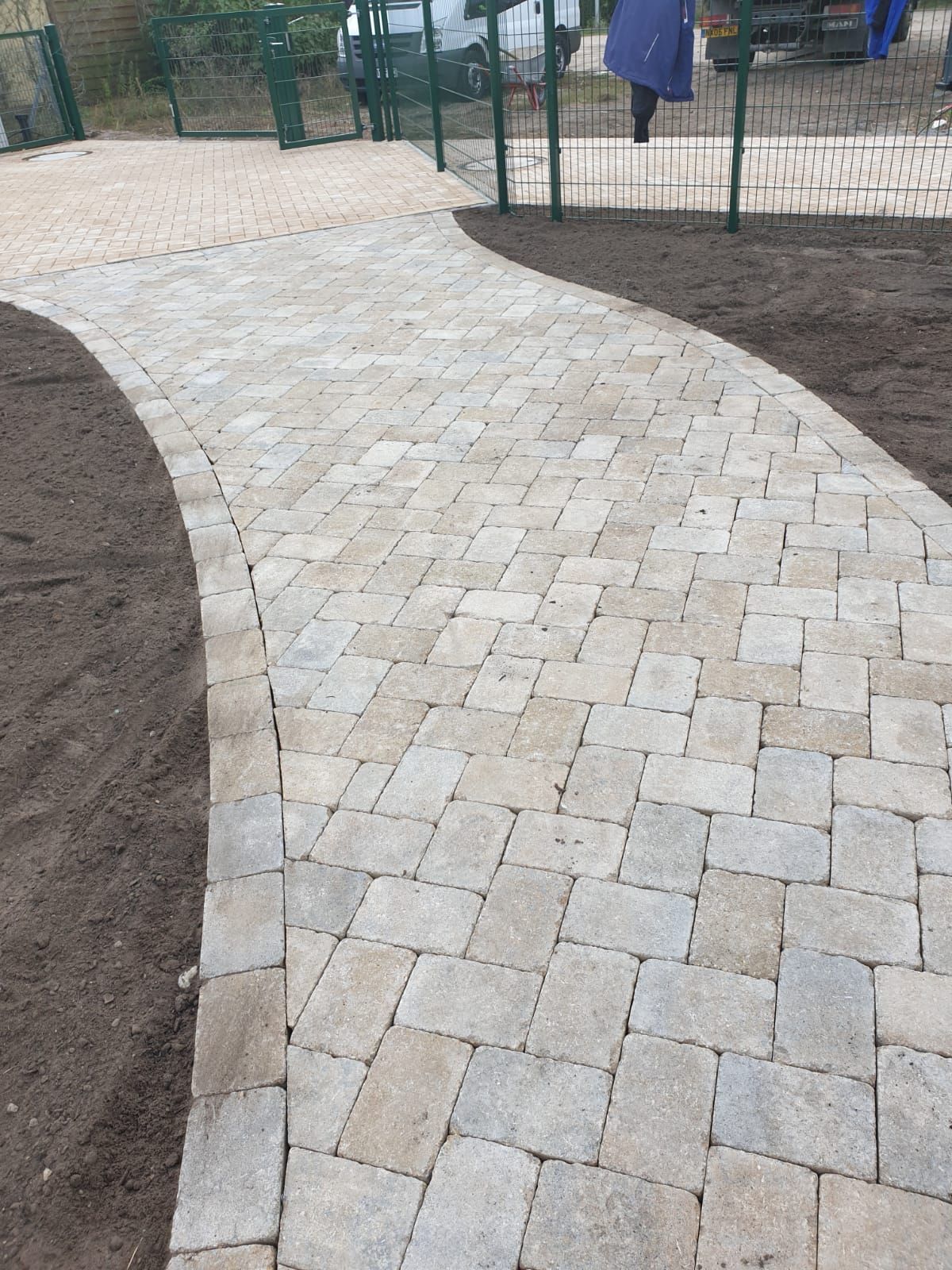 Brick pathway curves through a green space, leading toward a fence and figures in the background.