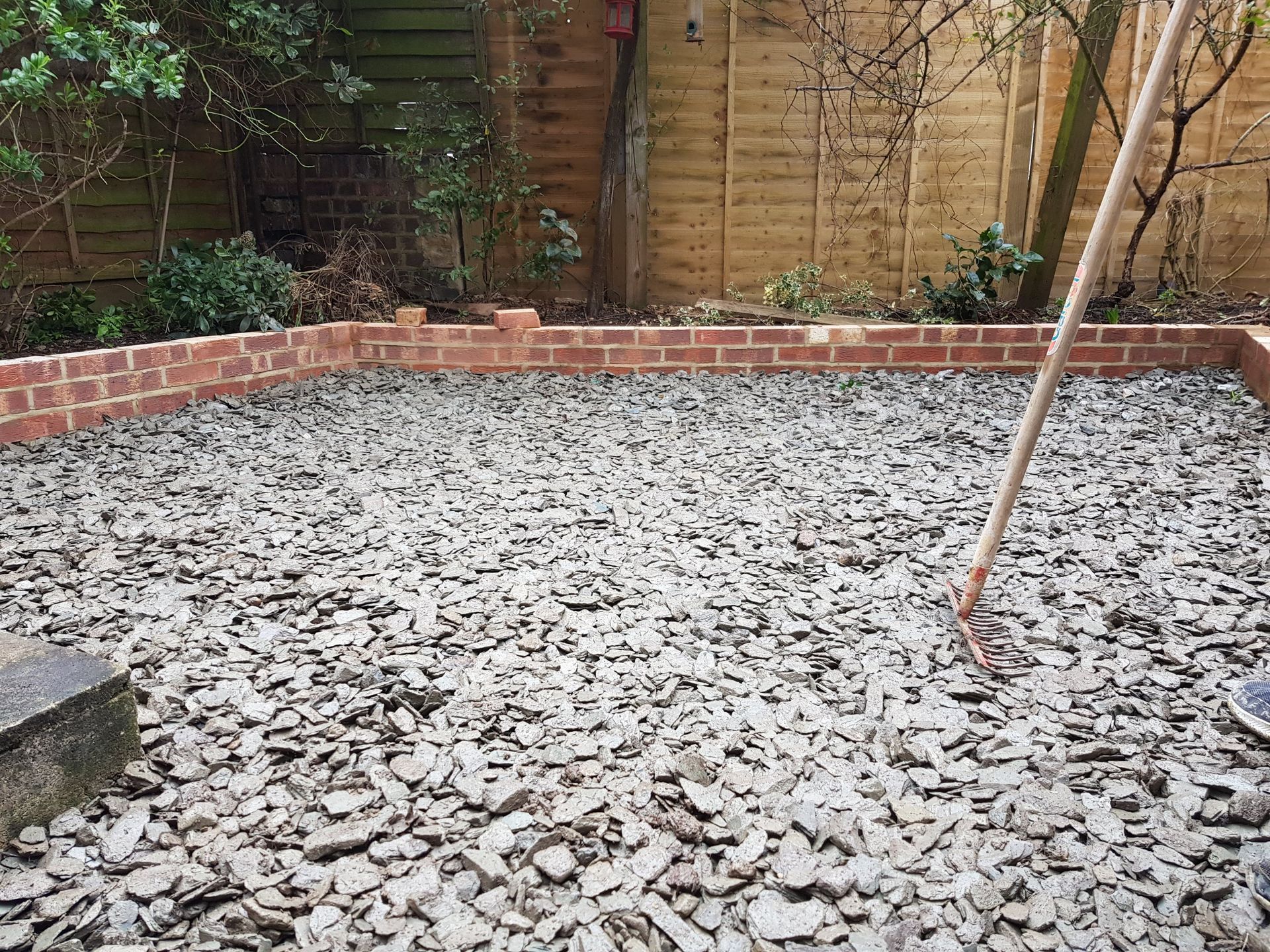 Gravel-covered patio with brick border, surrounded by wood fence and greenery. A tool rests in the center.