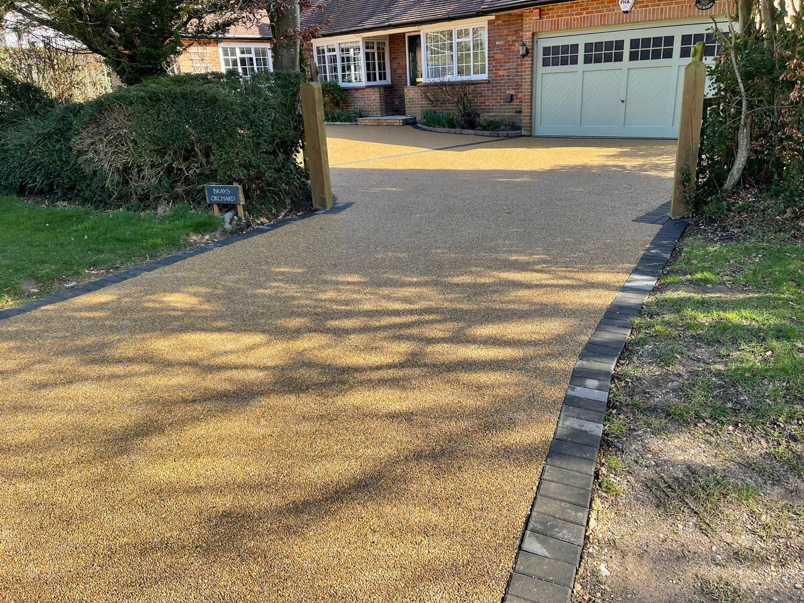 Gravel driveway leading to a brick house with a green garage door. Black border and wooden posts.