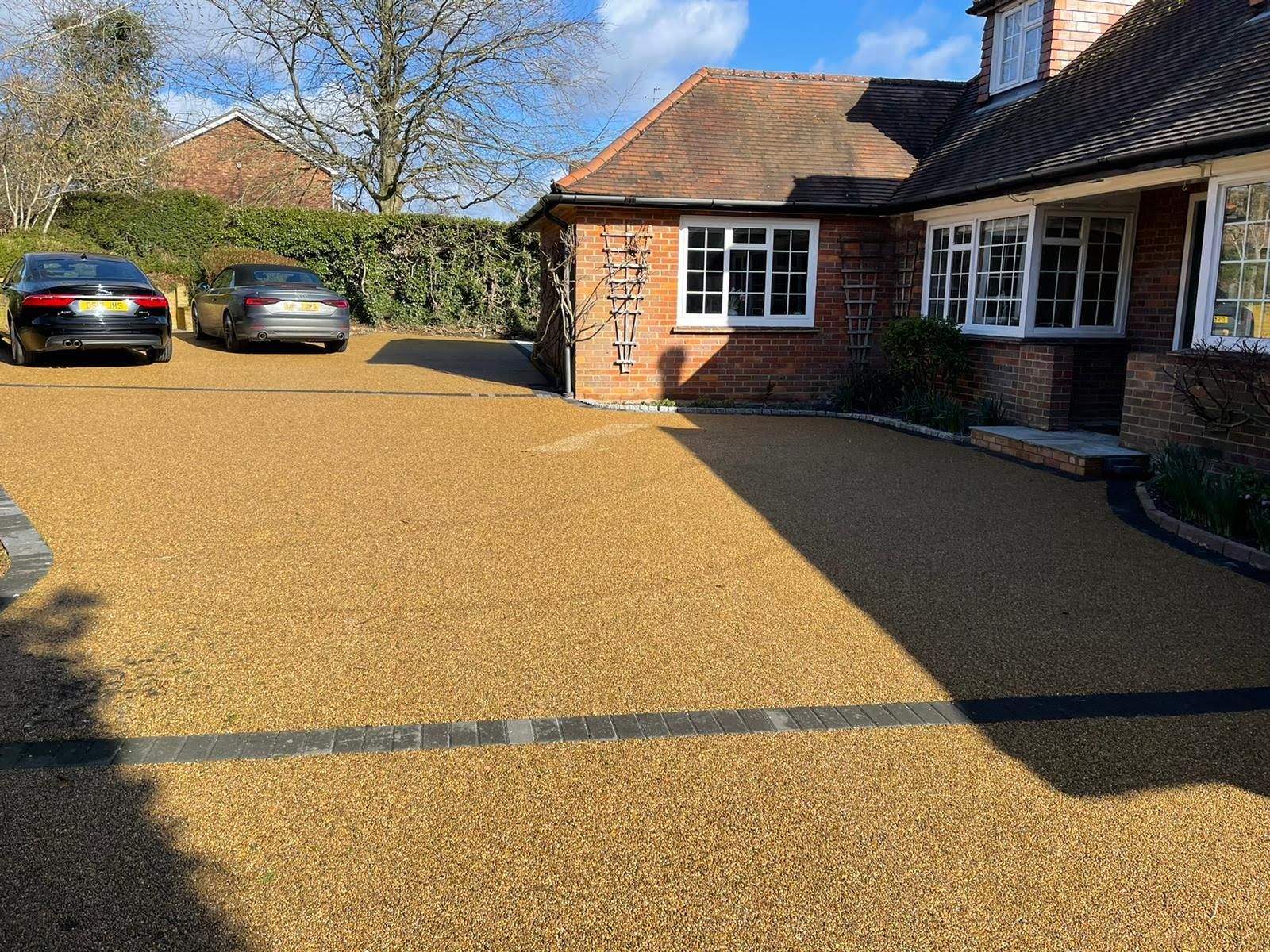 Tan gravel driveway with two cars parked in front of a brick house on a sunny day.